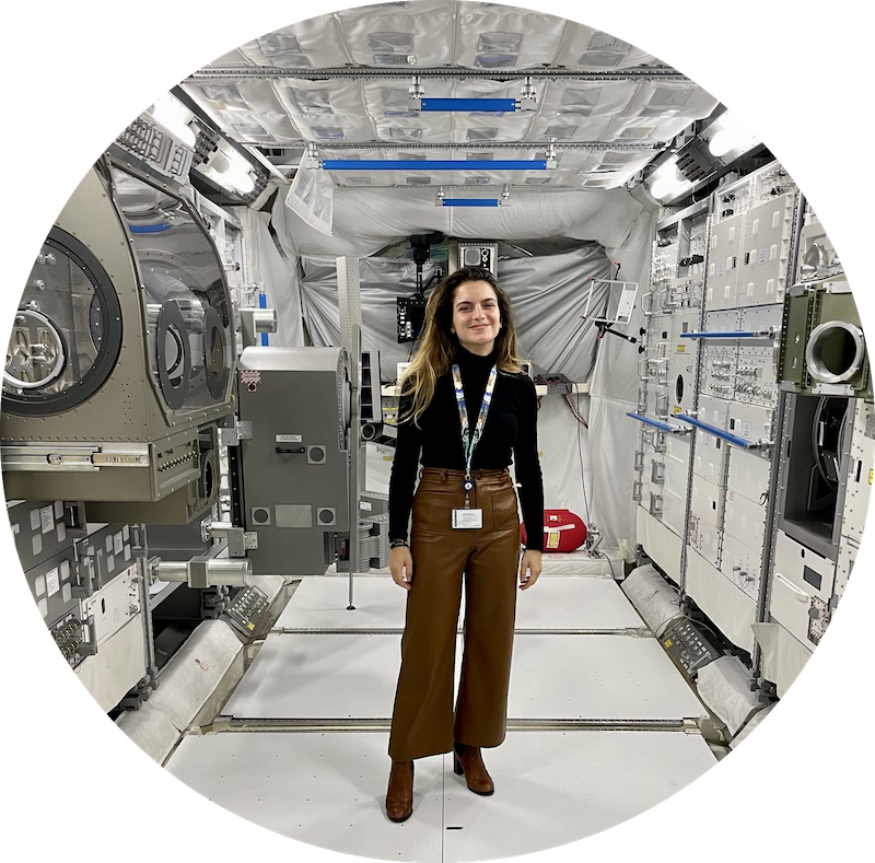 Smiling young woman with long hair standing in a cylindrical chamber between walls of complex machinery.