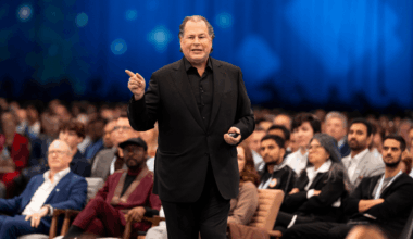 A man in a black suit standing around a crowd of people sitting, looking at him.
