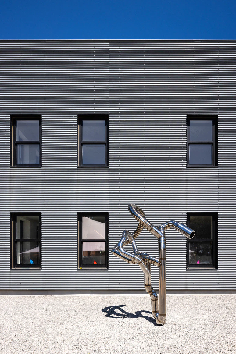 Modern building facade with striped pattern and unique metallic sculpture in foreground under clear blue sky.