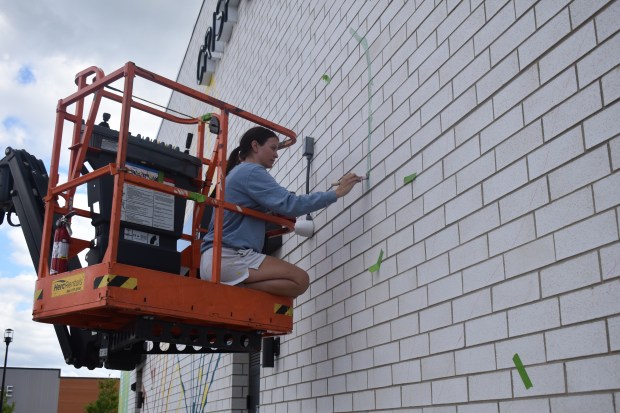 A boom lift allows artist Kailey Rorer to paint higher parts of a mural she's working on at Block 59 in Naperville. Bricks painted white are easier to work with as they don't need as many layers as do unpainted bricks, she said. (Steve Metsch/Naperville Sun)