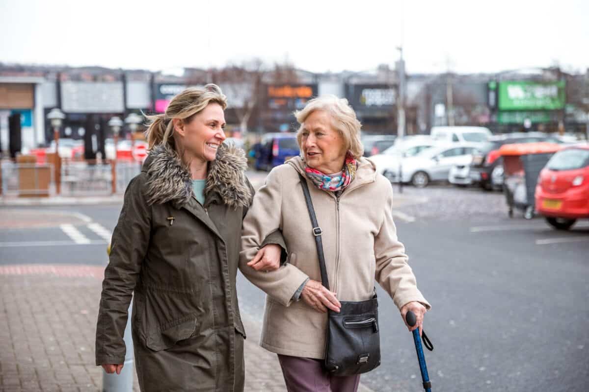 A mature woman help a senior woman out of a car as she takes her to the shops.