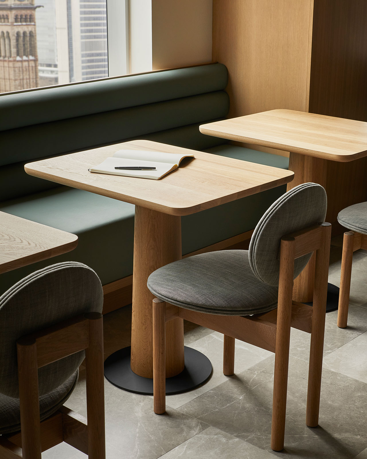 A wooden table with an open notebook and pen, surrounded by cushioned chairs and a built-in bench in a modern, well-lit room with large windows.