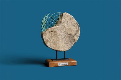 A studio shot of an artifact displayed on a small wooden base against a solid blue background. The artifact features a large, rough-textured stone disk with an uneven edge. Embedded or emerging from the upper-left edge of the stone is a green, weathered metal emblem resembling the United Nations logo, including an olive branch and a partial grid pattern. The stone structure is mounted on two short black metal pillars attached to a polished wooden stand, which has a small white label on the front that reads "EXHIBIT 1: The United Nations".