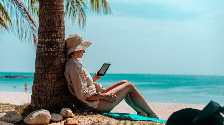 A woman sits on a beach against a palm tree reading from her Kindle.