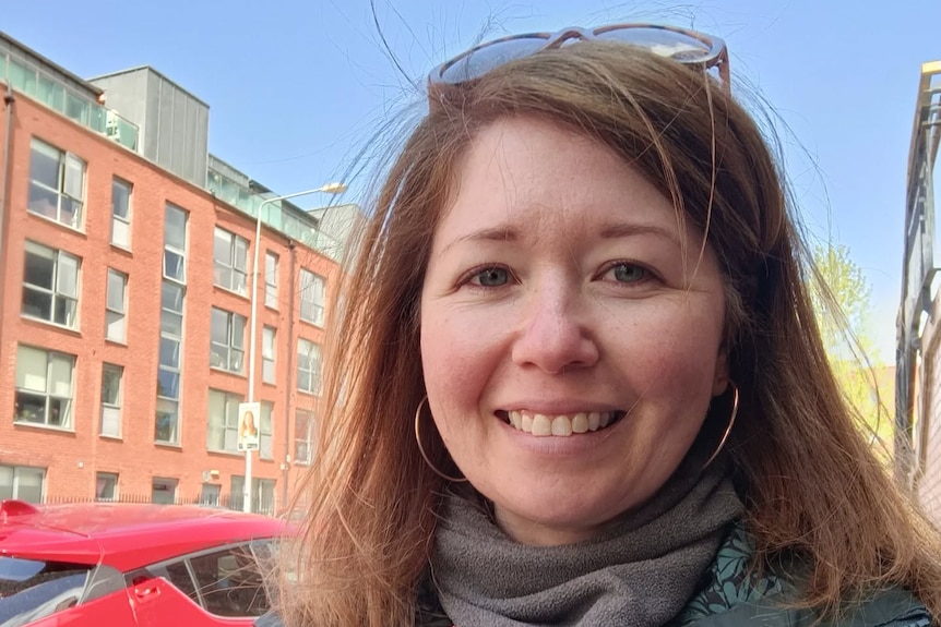 A white women in her 30s or 40s smiles at the camera on the street on a sunny day.