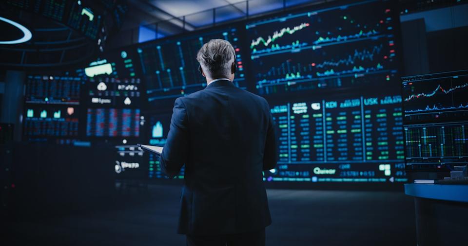 A man in a dark suit, seen from the back, stands in a dark, high-tech control room or trading floor. He faces multiple large, curved screens displaying glowing blue, green, and red financial charts, graphs, and stock data. He holds a white tablet in his left hand. The screens dominate the background, showing complex stock market information against a dark blue and black backdrop.