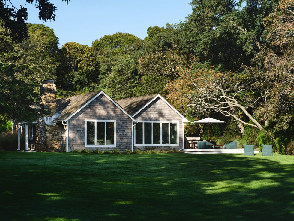 House with stone chimney in a lush green yard surrounded by trees.