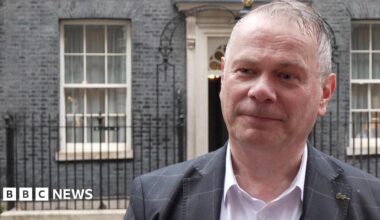 Corin Dalby, with receding grey hair and wearing a white shirt and grey suit with a lighter grey pattern, stands in the rain across the road from 10 Downing Street.