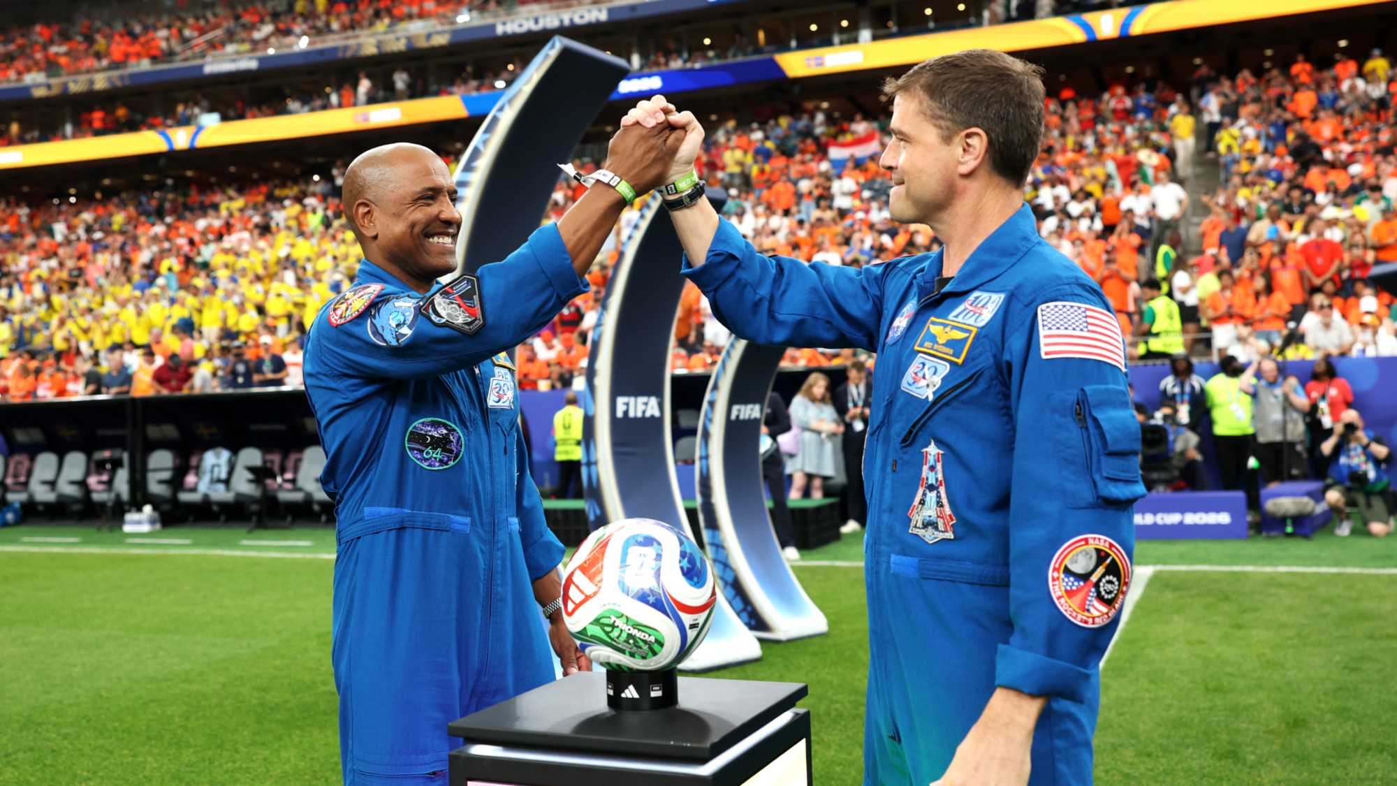 two men in blue flight suits smile and lock hands above a tricolor soccer ball on a grass field in a packed stadium