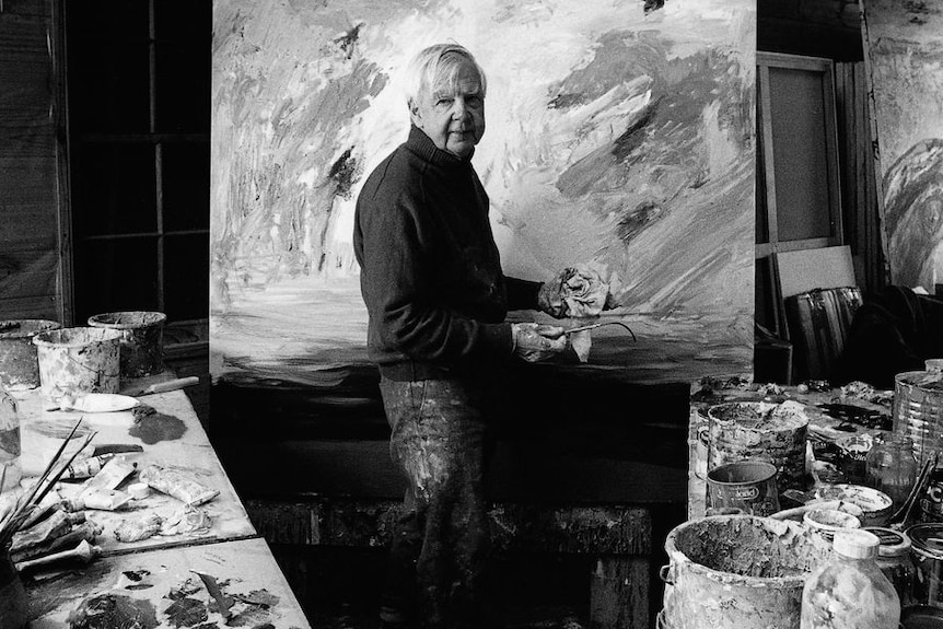A black and white image of an older man with white hair standing in a painting studio.