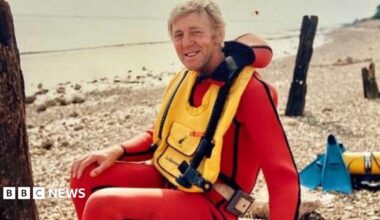 A man with fair hair sitting on a pebble beach. He has a red wetsuit on and a yellow life vest over the top. He is smiling as he looks into the camera, sideways facing.