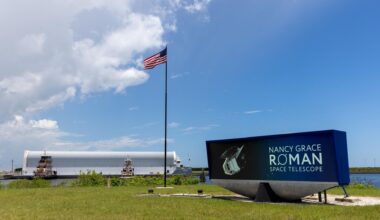 a long barge in the background arrives at a small offloading dock. In the foreground, and American flag in the center, and a digital display that says, "Nancy Grace Roman Space Telescope".