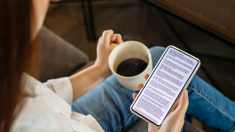A woman in white shirt and jeans is seen  from behind drinking a cup of black coffee and reading from her Kindle app.