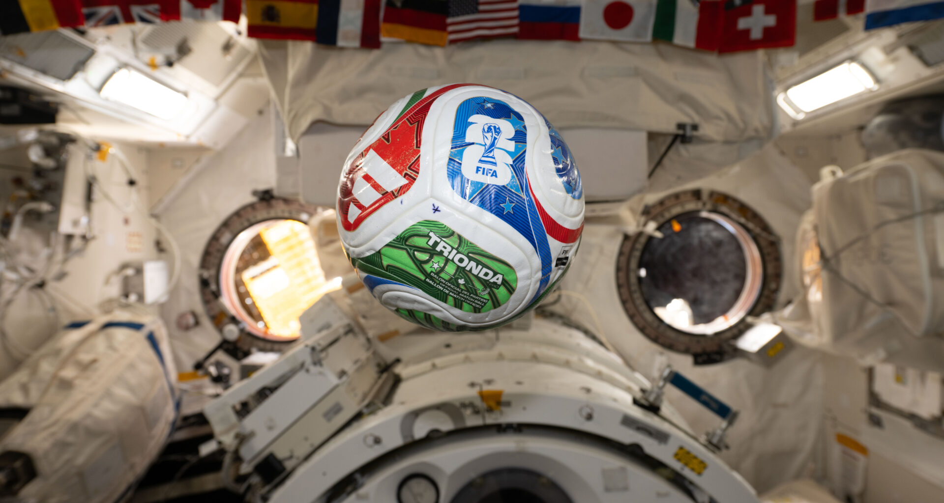 A white, red, blue, and green soccer ball floats inside the International Space Center. The FIFA logo is visible in the blue part of the ball facing the camera. The area in the background is mostly white, with a banner of country flags at the top of the photo.