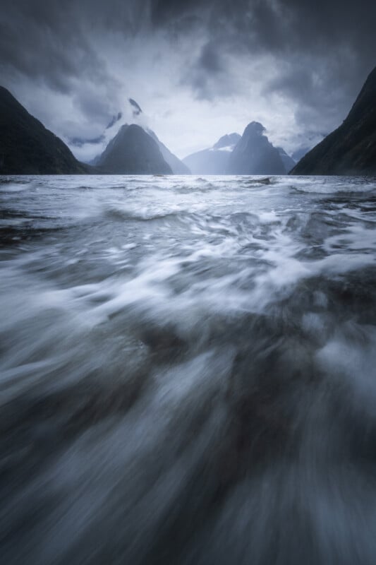 A dramatic, moody scene of fast-moving water flowing toward mountains shrouded in mist and clouds, under a dark, overcast sky. The perspective emphasizes the motion and power of the water in the foreground.