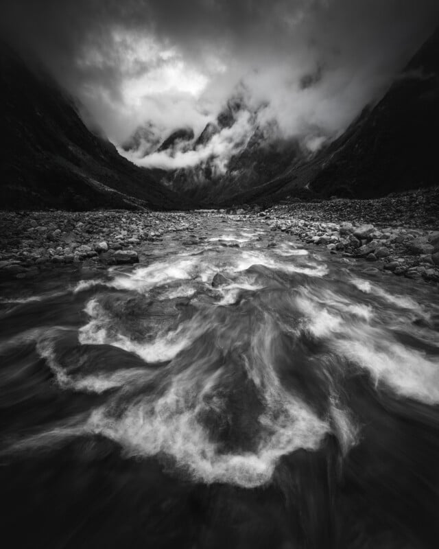 A dramatic black and white photo of a fast-flowing river with swirling currents, rocky banks, and misty mountains shrouded in clouds in the background under a moody sky.