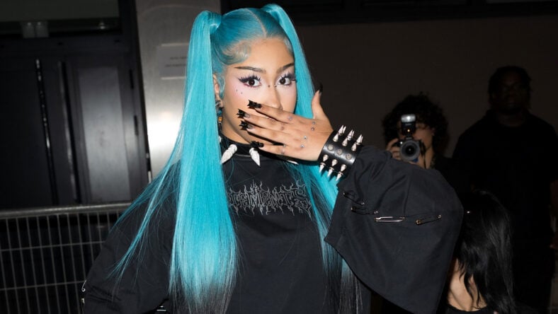 North West in a black grunge outfit with her blue hair in high pigtails. She holds her hands in front of her mouth while posing for a photo.