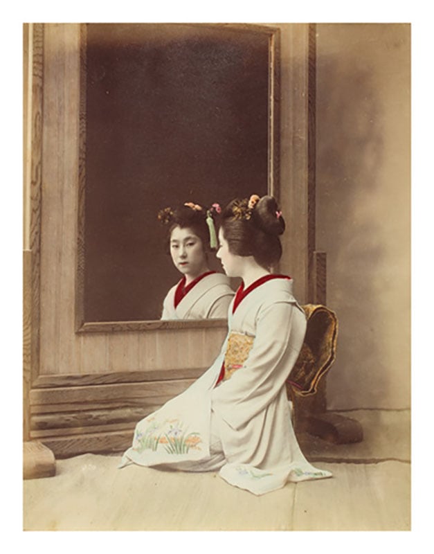 A woman in a traditional Japanese kimono kneels in front of a large wooden-framed mirror, looking at her reflection. Her hair is elaborately styled and decorated with ornaments. The background is neutral and simple.