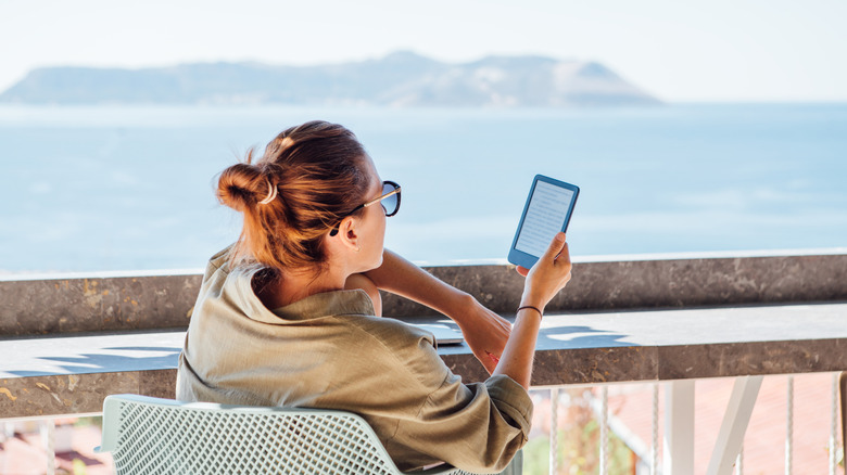A woman reads from her Kindle on a balcony overlooking the the sea, where an island can be seen in the distance.