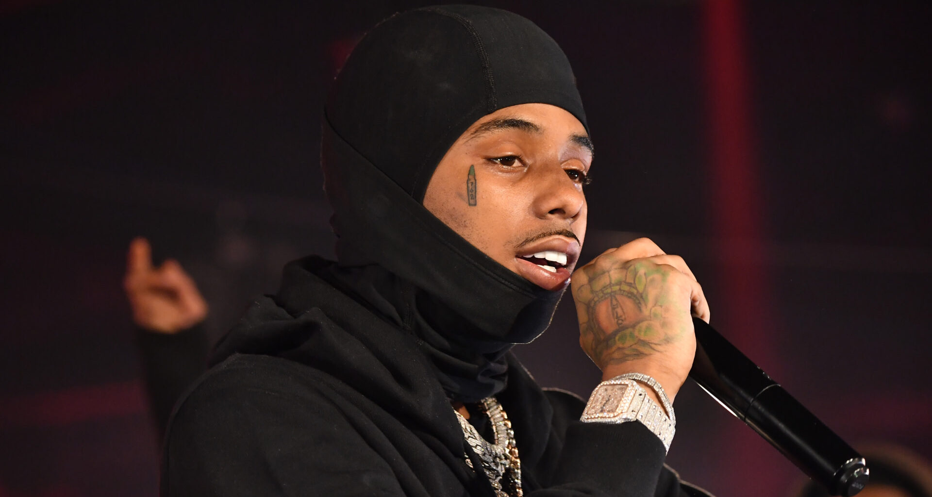 Pooh Shiesty wearing a black hood and holding a microphone performs on stage. They have a teardrop tattoo and visible jewelry.
