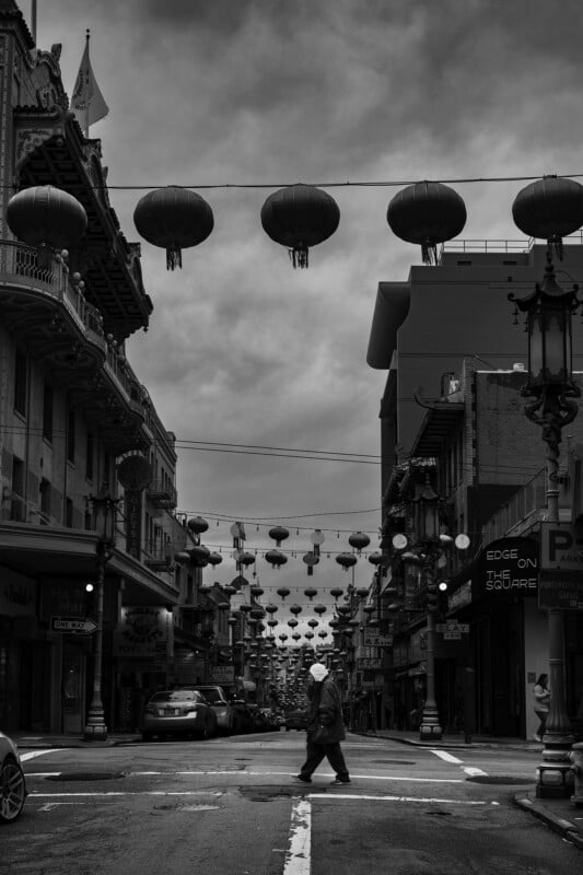A person crosses a street in Chinatown, surrounded by buildings and rows of hanging lanterns under a cloudy sky; the scene is in black and white.