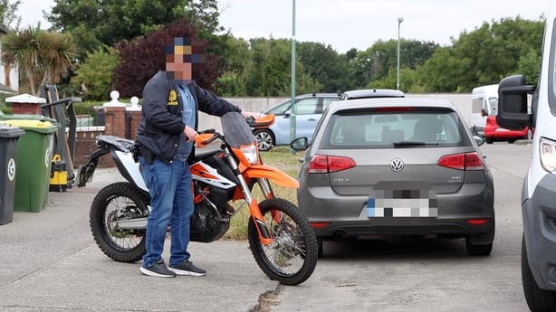 A man with a blurred face in a Garda jacket holds a scrambler bike