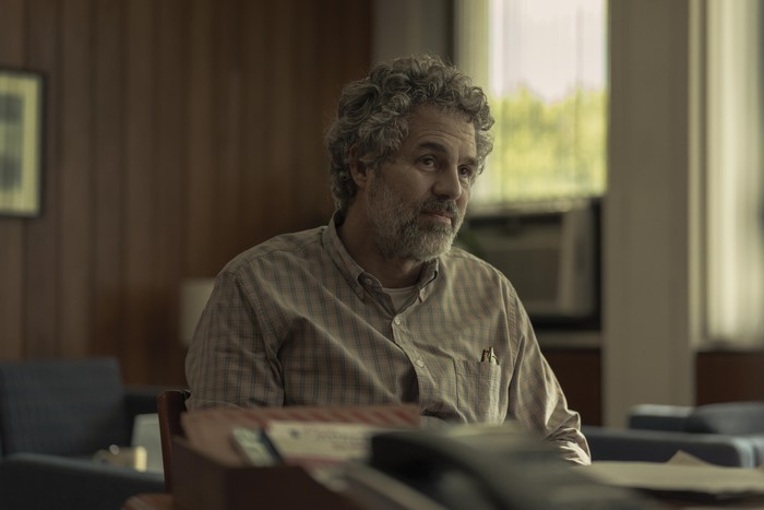 Task Mark Ruffalo in Task, wearing a shirt and sat behind a desk.