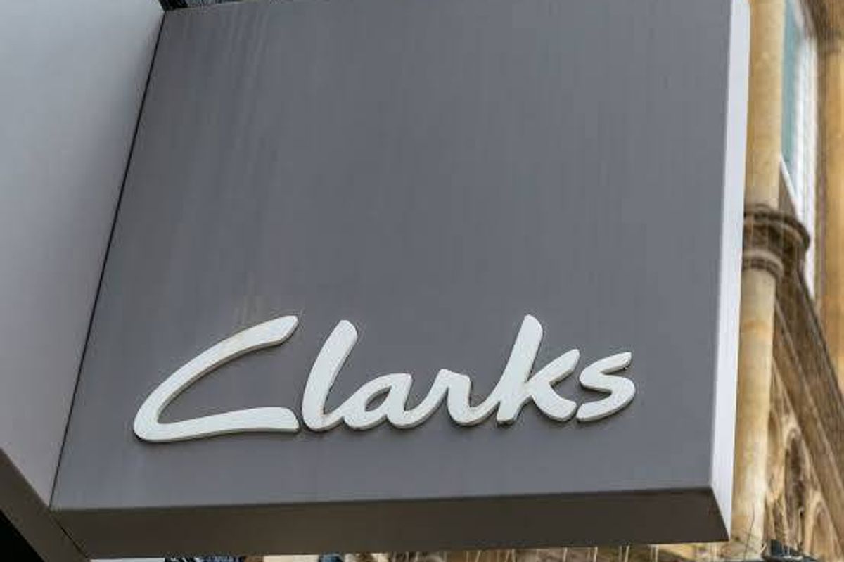 An image displays a storefront sign, prominently featuring the word "Clarks" in white text on a gray background. The sign is affixed to the side of a building, with a blue banner partially visible below.