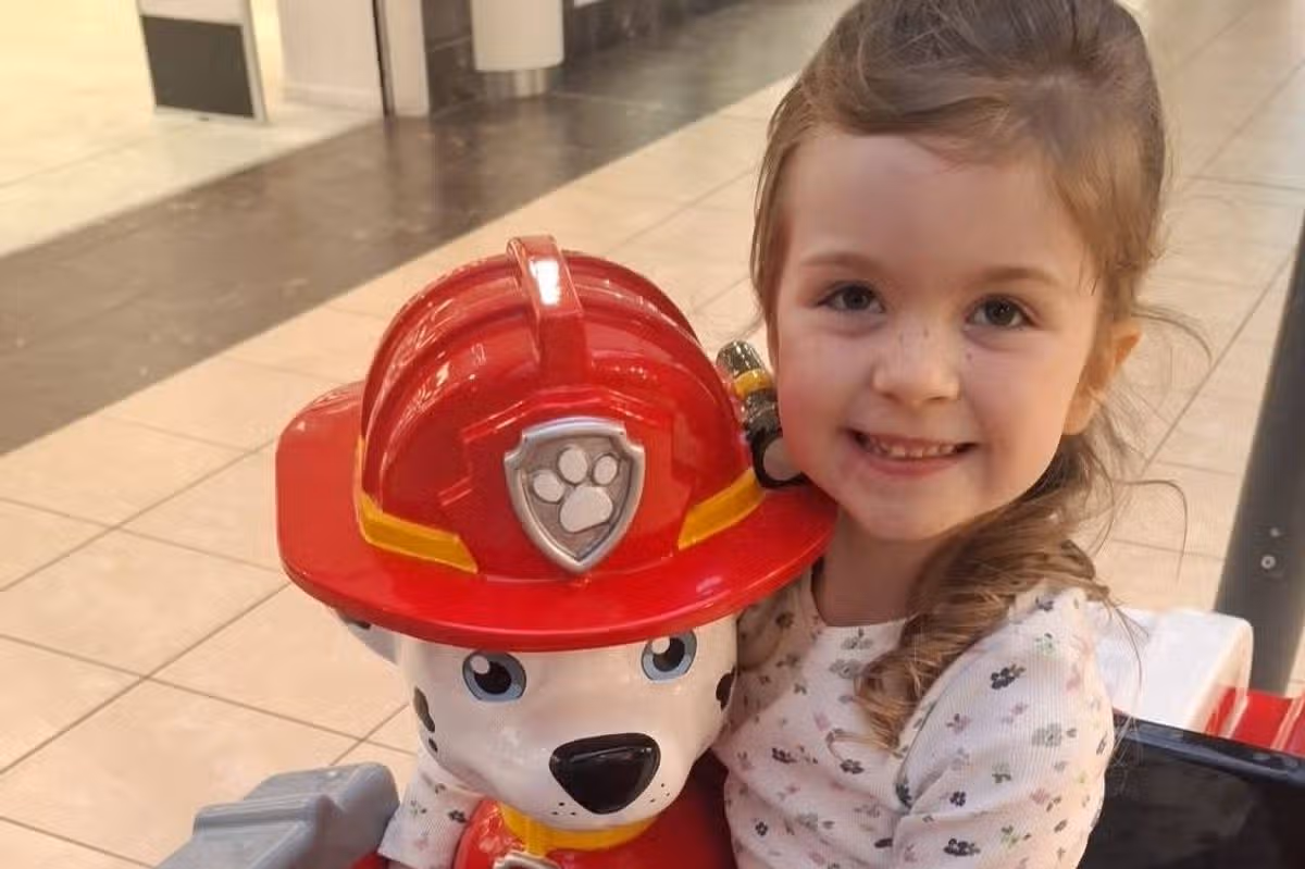 A young child wearing white pajamas is seated with a toy figurine of a firefighter's dog, holding the dog's helmet and smiling. The setting appears to be an indoor public space with tiled flooring and visible lighting overhead.