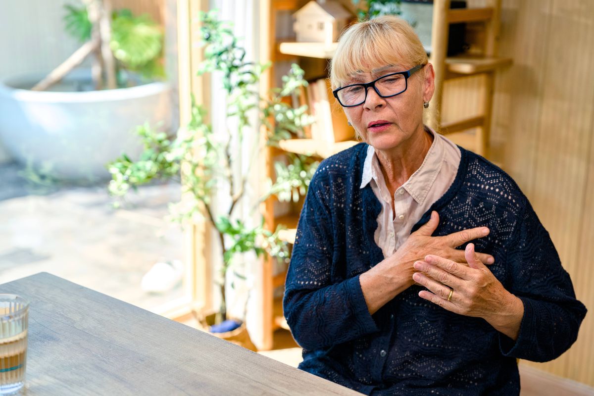 Senior woman sitting at a table feeling sudden chest pain and shortness of breath, indicating a potential heart attack or other cardiovascular health issues at home