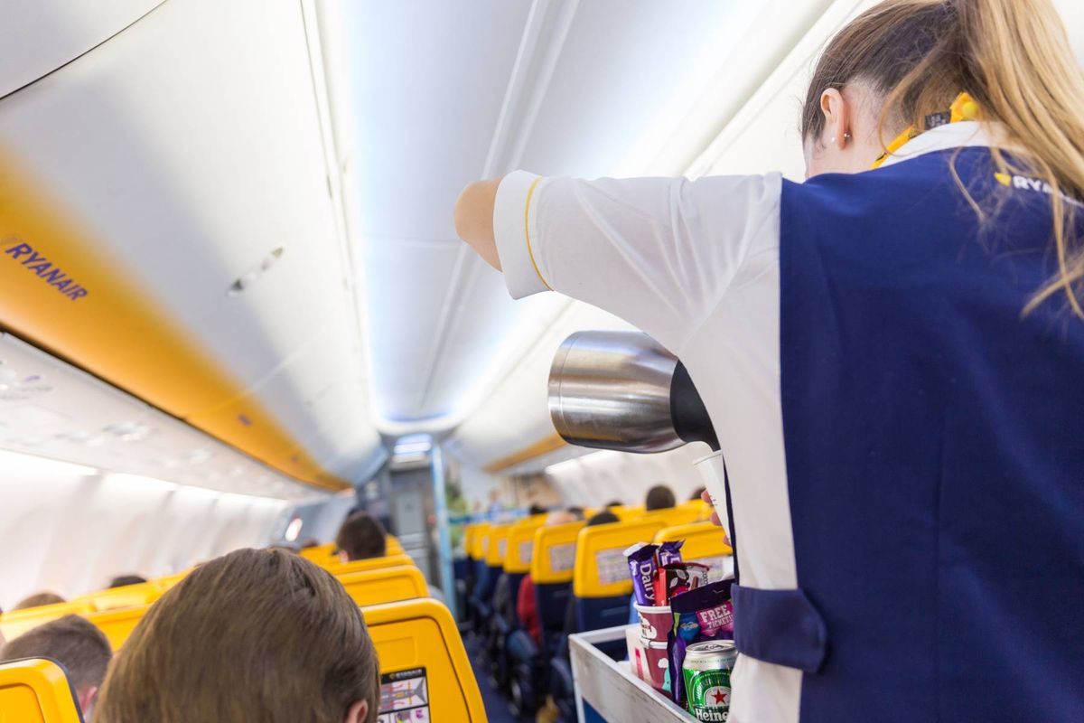 A cabin crew member is seen attending to the needs of passengers aboard an aircraft. The interior of the plane is visible, featuring yellow seats and a white ceiling. The crew member is dressed in a blue and white uniform and is reaching over the seats to assist a passenger.