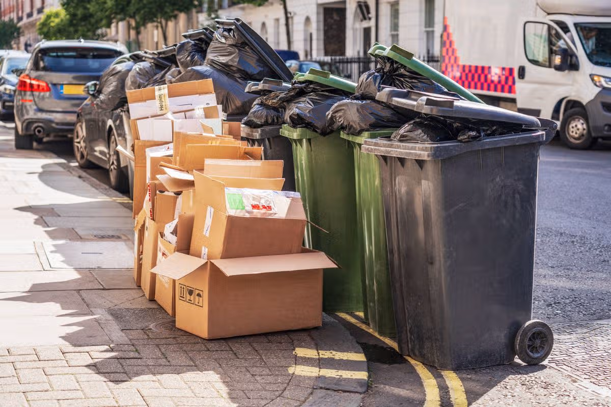 Several black and green wheel bins full of rubbish bags, alongside a collection of cardboard boxes for recycling on a residential street in London.