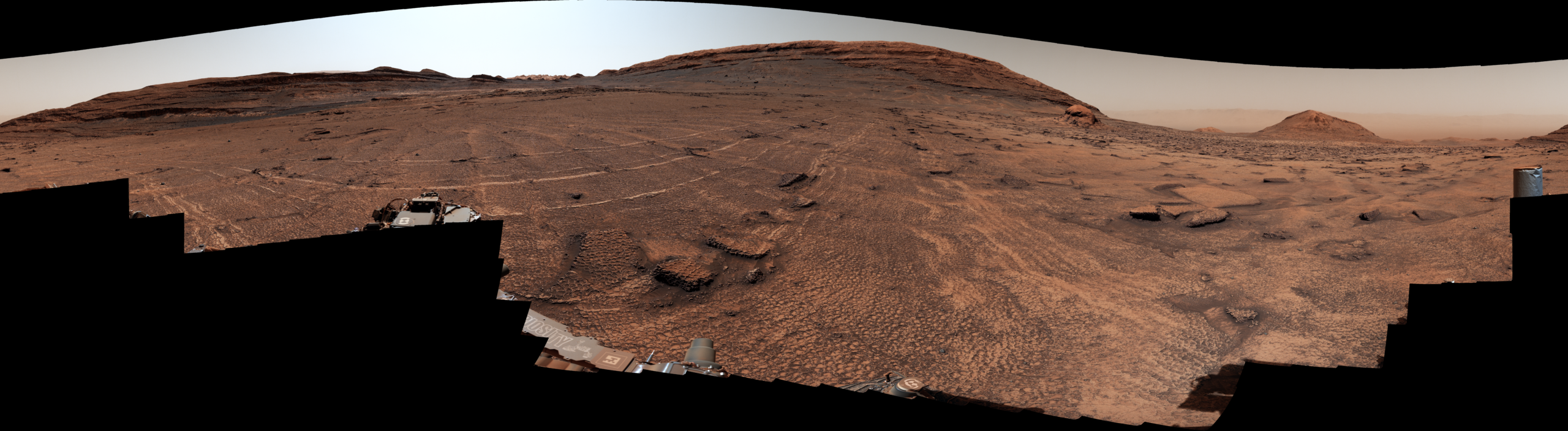 A panoramic view of a sweeping Martian plain covered in patterned fractures leading toward distant hills showcases the terrain explored by NASA's Curiosity rover to investigate the planet's past environment.