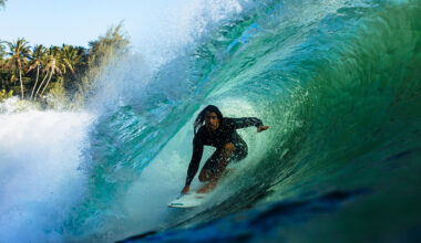 A surfer in a black wetsuit crouches on a surfboard inside a large curling turquoise wave as the waves break over him. Palm trees are visible in the background.