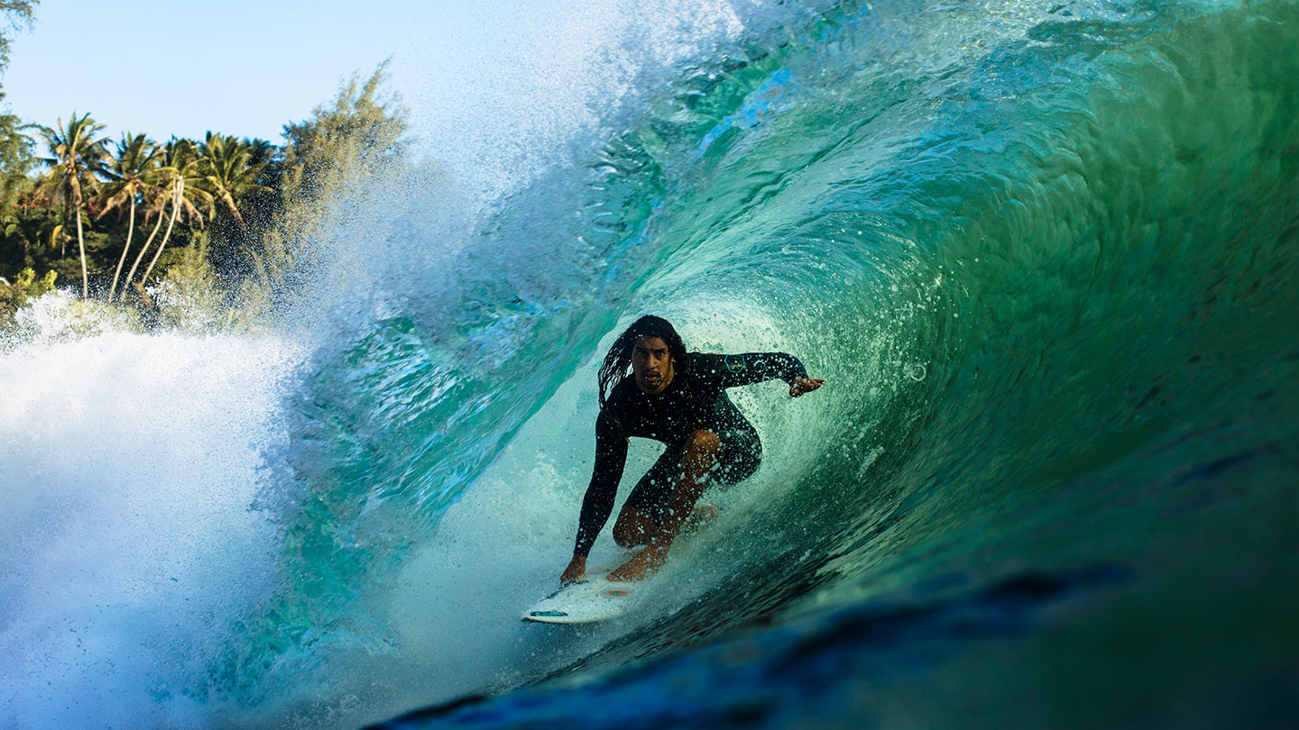 A surfer in a black wetsuit crouches on a surfboard inside a large curling turquoise wave as the waves break over him. Palm trees are visible in the background.
