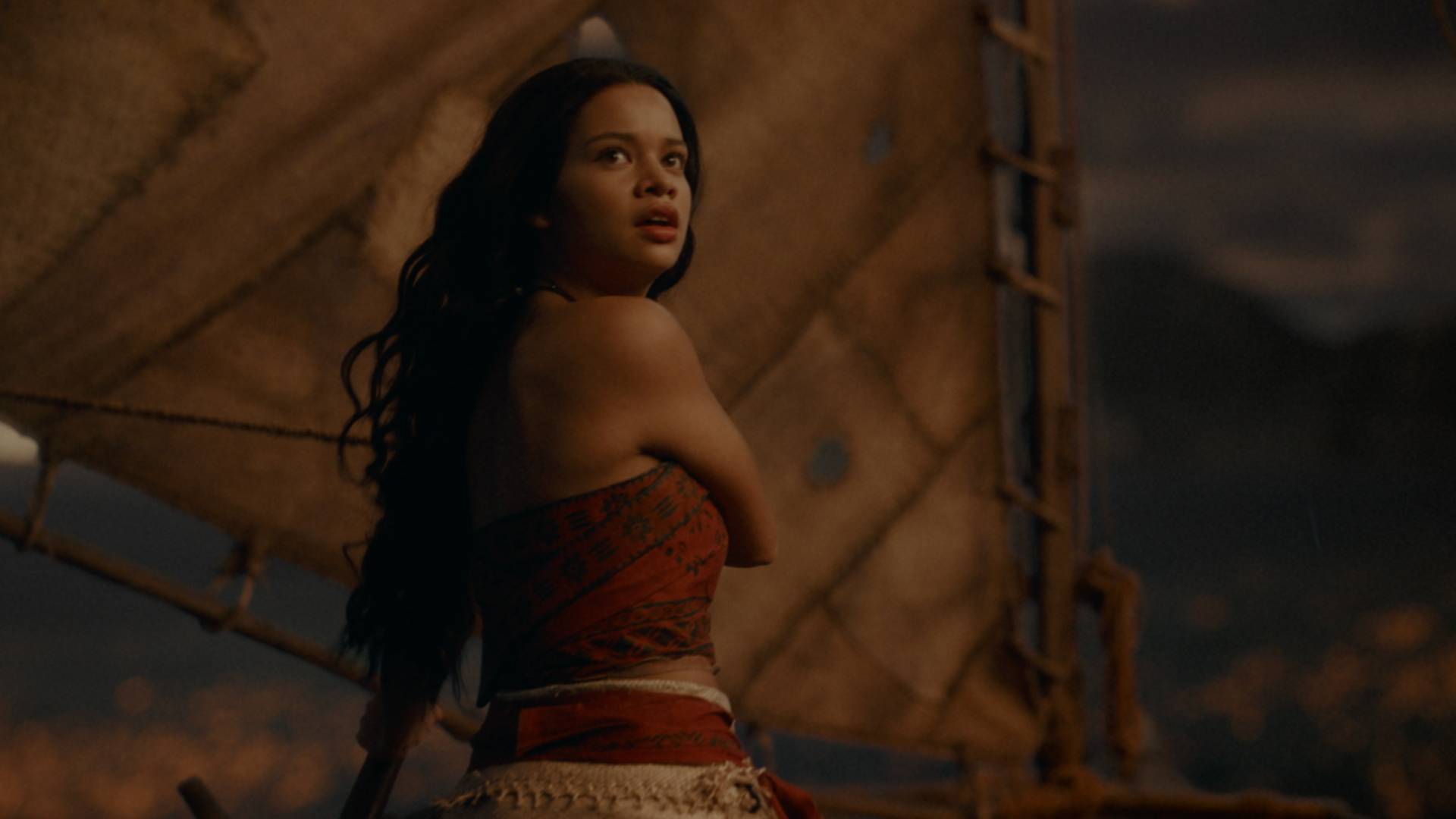 A live-action Moana stands on a sailboat, looking over her shoulder with an alarmed expression against a backdrop of sails and a dim, warm-toned horizon.