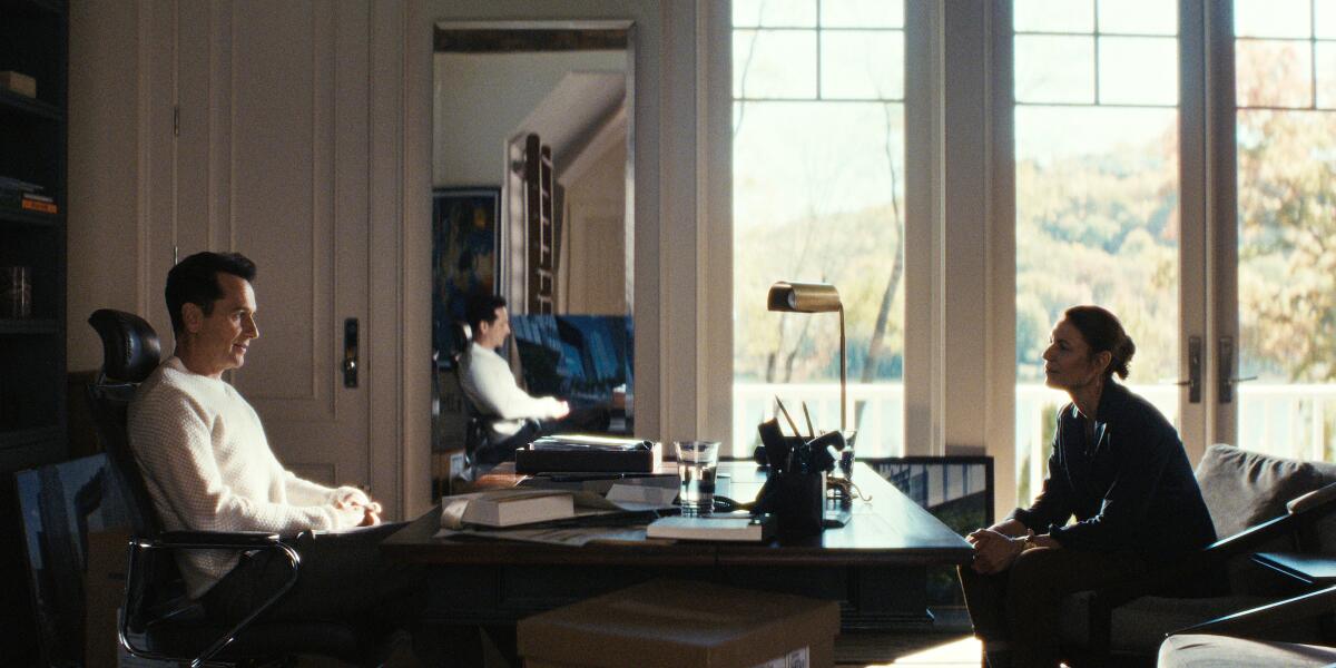 A man in an office chair sitting behind a desk looking at a woman seated opposite of him.
