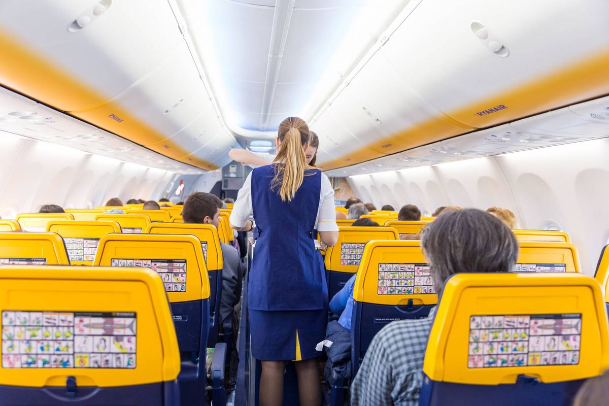 Interior view of a commercial airplane with rows of blue and yellow seats, and a person in a blue uniform standing in the aisle, likely a flight attendant.