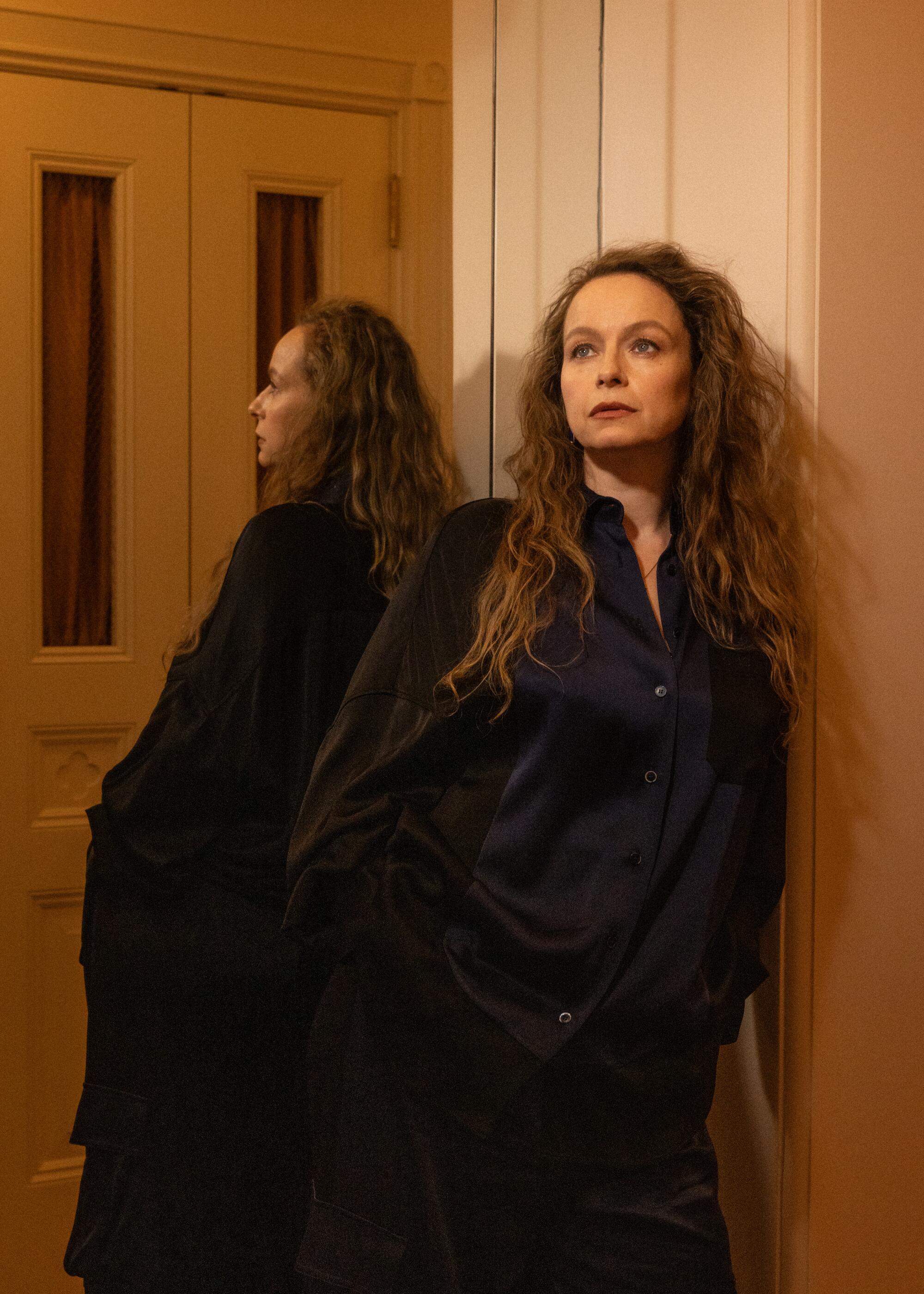 A woman in a blazer stands by a mirror in a hotel room.