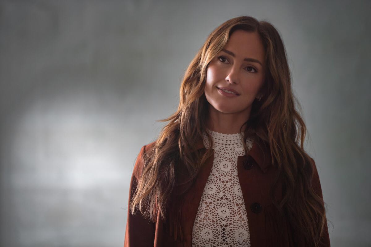 A brunette woman, wearing a white lace top and a brown suede jacket, stares ahead
