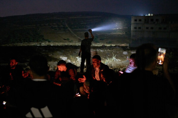 A Palestinian scans a valley with a flashlight as people gather around a bonfire during a volunteer night watch to help prevent Israeli settler attacks in the West Bank town of Sinjil, Monday, July 27, 2026. (AP Photo/Leo Correa)