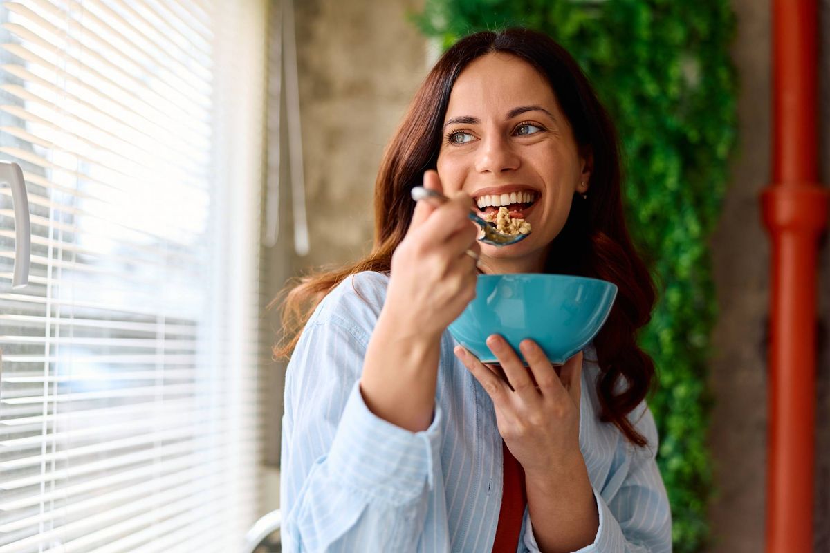 Smiling mid adult woman enjoying a nutritious breakfast bowl with cereal and fruit, looking out of the window during a bright morning at home, embracing a healthy lifestyle