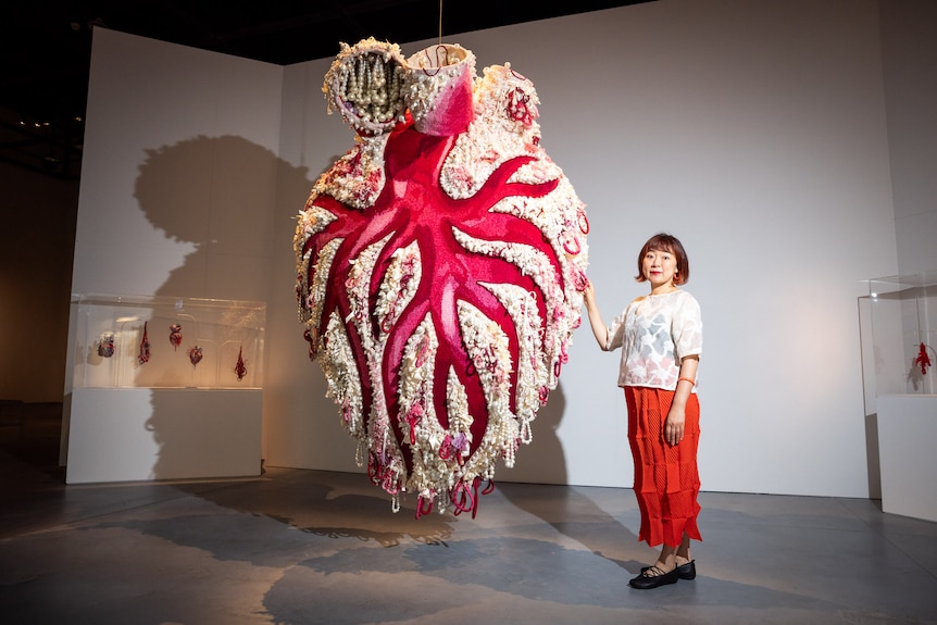 A Japanese Australian woman in a gallery stands beside a large textile sculpture of a heart, which features beads and ribbon.