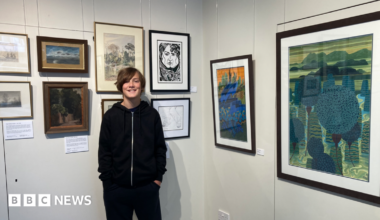 Valya Constable smiles for the camera next to his framed artwork in the exhibition