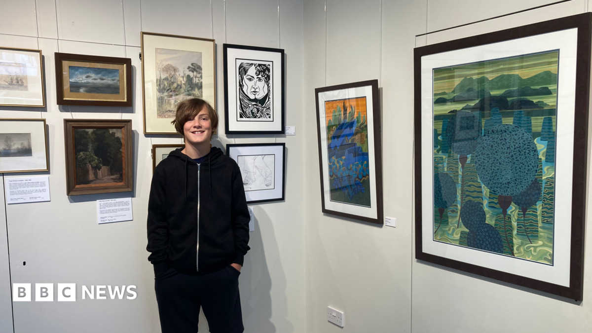 Valya Constable smiles for the camera next to his framed artwork in the exhibition