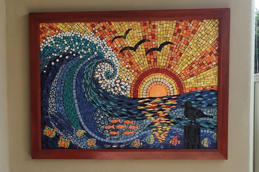 A mosaic of a sunset over a wave. It hangs on a wall that leads to a backyard.