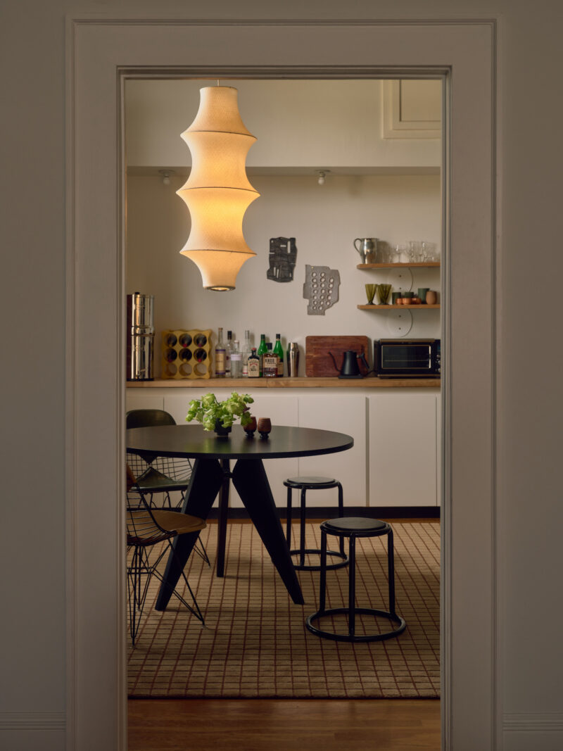 A modern dining area with a round black table, four chairs, a sculptural hanging lamp, and a kitchen with shelves and various items in the background.