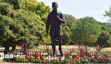 A life-size, grey sculpture of Brian Clough. He appears to be walking with his football boots slung over his shoulder by their laces. The figure has short hair and is wearing shorts and a long sleeve top. The statue is surrounded by red and white flowers and there are trees in the background.