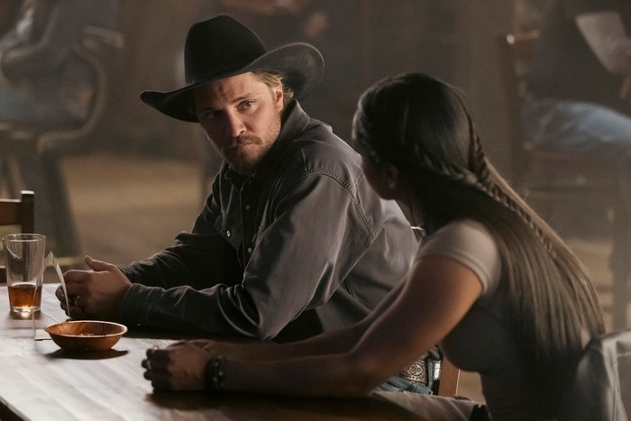 Family Business Luke Grimes as Kayce Dutton in Marshals, sat at a bar, wearing a shirt and a stetson and talking to a woman.