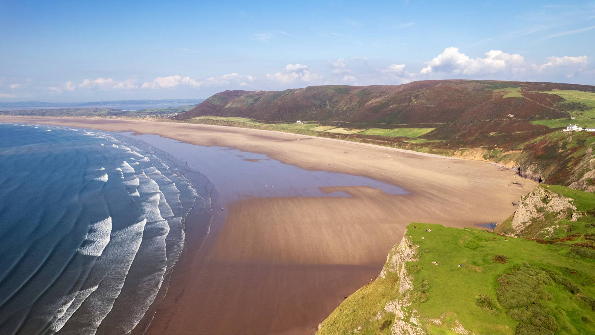 A wide sandy beach stretches along the coastline with gentle waves lapping the shore, bordered by grassy green cliffs and rolling hills under a blue sky with scattered clouds.